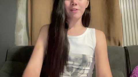 Video of a young woman with long, straight black hair, fair skin, wearing a white tank top with a black graphic, sitting on a gray couch against a beige wall.
