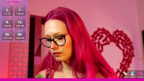 Media: A video of a fair-skinned woman with long, vibrant pink hair, wearing black-framed glasses, and a tattooed arm. She stands in a room with pink and purple hues, featuring a red heart-shaped wall decoration.