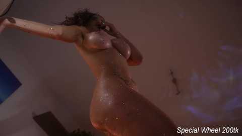 Video of a nude, dark-skinned woman with wet, shiny skin, standing in a dimly lit room. She has a curvy physique, medium-sized breasts, and is posing with one arm above her head. The background includes a blue wall and a wooden shelf.