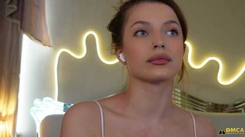 A video of a young Caucasian woman with fair skin, brown hair in a bun, wearing white earphones, and a white spaghetti-strap top. She gazes thoughtfully at a glowing neon wave mural in the background.