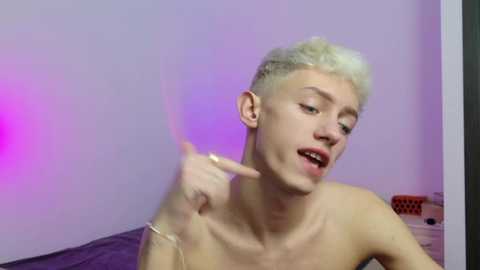 Video of a young, fair-skinned, shirtless man with platinum blonde hair, making a \"rock on\" gesture, in a dimly lit room with purple and pink ambient lighting.