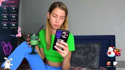Video of a blonde woman with fair skin in a green crop top and blue leggings, sipping a green drink, in a dimly lit room with a dark headboard, pink decorations, and a digital interface displaying streaming statistics.