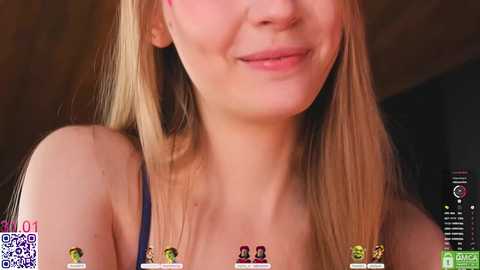 Video of a smiling young woman with long blonde hair, wearing a blue spaghetti strap top, against a blurred dark background.