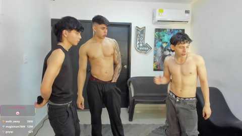 Video of three shirtless, muscular young men in a modern, minimalist room. One wears black pants, another has tattoos, and the third has a distressed look.