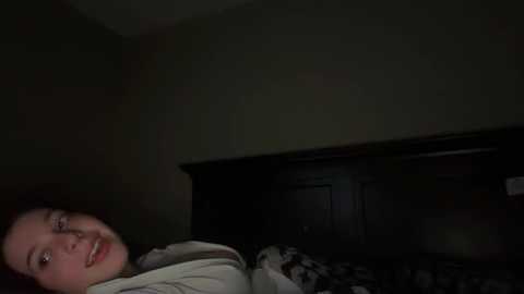 Video of a young woman lying on a dark wooden bed with a patterned blanket, partially covered by a white sheet. Her face is partially visible, showing a calm expression. The room is dimly lit, creating a moody atmosphere.