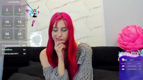 Video of a young woman with bright red hair, wearing a gray knitted sweater, sitting on a black couch, in a modern room with a pink flower, digital screen displaying social media notifications.