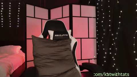 Video of a black gaming chair with a gray pillow covering its head, in a dimly lit room with a black folding screen and string lights.