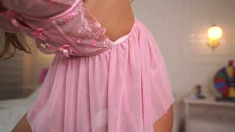 Video of a woman in a pink, sheer, lace-trimmed nightgown, with a close-up view of her back and shoulders, showing the delicate fabric. The background features a beige wall and a colorful, abstract painting.