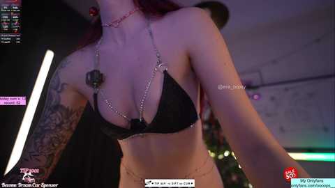 A video of a light-skinned woman with long red hair, wearing a black bikini top with a chain design, tattoos on her arms, in a dimly lit room.