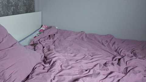 A video of a messy, unmade bed with pink sheets and a white headboard against a gray, textured wall. The bedding is wrinkled and disheveled, with a pillow lying on the floor beside the bed. The room appears sparsely decorated and dimly lit.