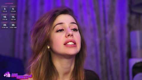 Video of a young Caucasian woman with light skin, straight brown hair, and subtle makeup, speaking into a microphone. Background shows purple lighting and a blurred, indistinct scene.