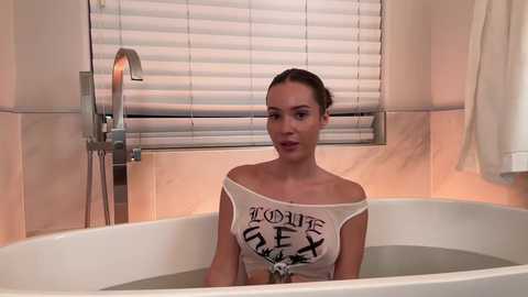 Media: Video of a light-skinned woman with dark hair in a white off-shoulder crop top, sitting in a bathtub with partially visible water, in a bathroom with beige tiles, a white towel, and a window with blinds.