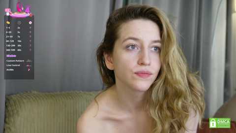 Video of a fair-skinned, blue-eyed woman with wavy blonde hair, wearing a light-colored top, seated on a green couch with beige curtains in the background.