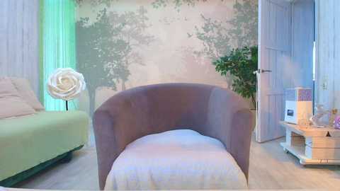 A video of a modern, minimalist bedroom featuring a curved, dark gray armchair, light green sofa, white wall mural with tree design, and a large leafy plant on a light wooden floor.