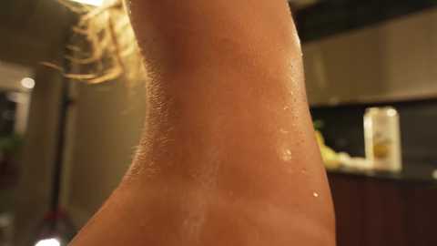 A video showing a close-up of a woman's wet, glistening arm in a kitchen setting. The background features a blurred stove, cabinets, and a bottle of cooking oil.