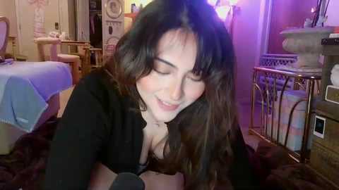 A video of a young woman with long, wavy brown hair and fair skin, wearing a black top, lying on a bed in a cozy room with a pink light, laundry baskets, and a cat.