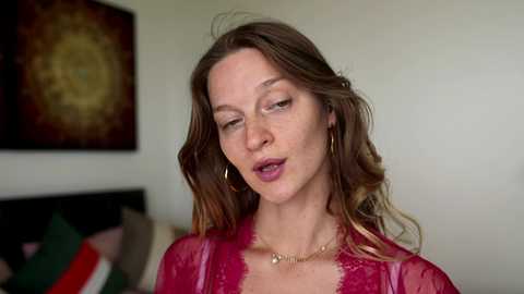 Video of a Caucasian woman with light skin, long brown hair, wearing a sheer, magenta lace top and gold hoop earrings, in a modern living room with abstract art on the wall.