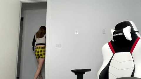 A video shows a young woman in a yellow plaid skirt and white crop top peeking into a gaming room with a white and black gaming chair and monitor.