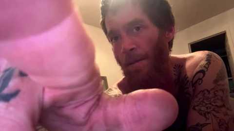 Video of a tattooed man with a beard and a large, erect penis in front of him, in a dimly lit room with beige walls.