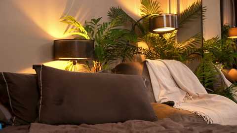 Video of a cozy, well-lit bedroom featuring a bed with brown and white pillows, a beige throw blanket, and lush green plants in the background.