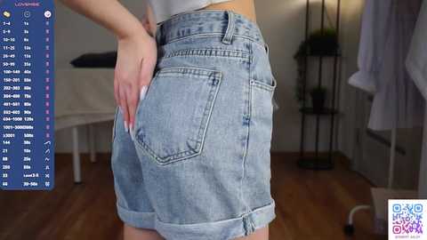 Media: Video of a woman wearing light blue denim shorts with a pocket, standing indoors with a white background, showing her backside and right hand on her hip.