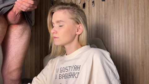 Video of a blonde, fair-skinned woman in a white robe with Russian text, seated, eyes closed, looking serene. A man's exposed penis is visible in the background.