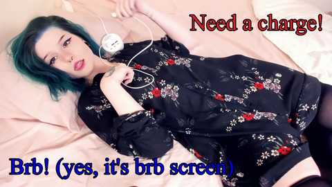 Video of a young woman with teal hair, lying on a bed, holding a white charger, wearing black floral pajamas, and red lipstick. Text overlays read \"Briol (yes, it's in beta screen)\" in blue and \"Need a charge!\" in red.