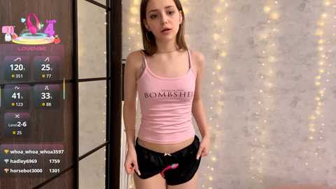 Video of a young, fair-skinned woman with light brown hair, wearing a pink tank top and black shorts, standing in a room with a wooden door, a digital display showing weight and age statistics, and string lights in the background.