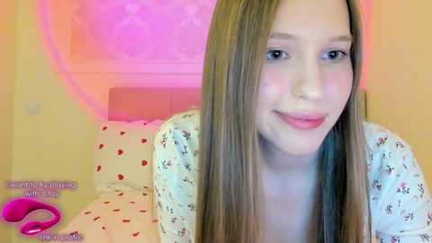 Video of a young Caucasian woman with long blonde hair, wearing a floral top, smiling. Background shows a pink wall, bed with heart-patterned sheets, and a pink dildo on the floor.