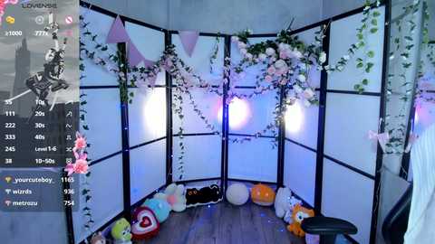 A video of a cozy, minimalist room adorned with white walls, black frames, and pastel pink flowers. The floor is covered with plush toys, including a large, smiling plush character. The room is illuminated by soft, pastel-colored lights.