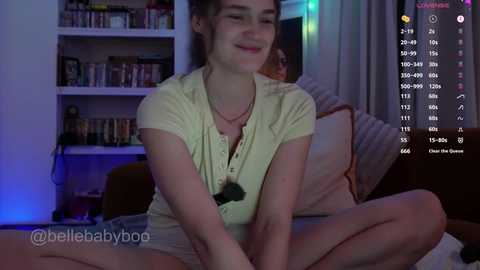 Video of a smiling young woman with light skin and brown hair, wearing a yellow blouse, sitting cross-legged on a bed, with bookshelves and a TV in the background.