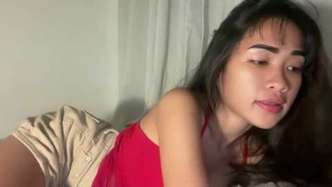 Video of a young Asian woman with long black hair, wearing a red tank top and beige shorts, lying on her stomach, looking slightly to the side, in a dimly lit room with white curtains.