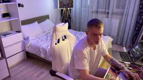 Video of a young, tattooed, white man with short, blonde hair, playing video games on a laptop, sitting in a white gaming chair, in a modern, well-lit bedroom with a bed, shelves, and a window.