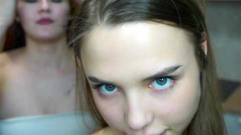 Video of a close-up of a young woman with fair skin, blue eyes, and straight brown hair, looking intently forward. Another woman with fair skin and long brown hair is blurred in the background.
