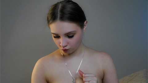 Video of a young, fair-skinned woman with brown hair tied back, wearing a thin silver necklace, topless, with a blurred background.