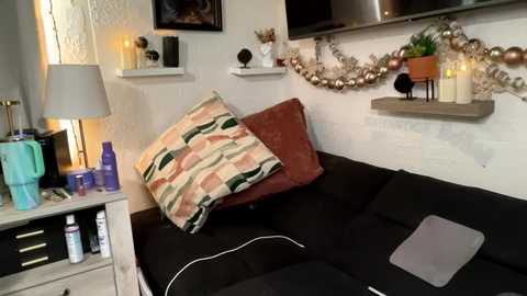 Video of a cozy, modern living room with a black sofa, patterned pillows, white shelves, candles, plants, and a TV mounted on the wall.