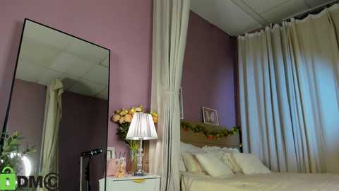 Video of a cozy bedroom with mauve walls, white curtains, a large mirror, a bed with white linens, a nightstand with a lamp, and a bouquet of flowers.