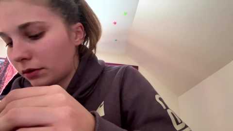 A video of a young woman with light skin and brown hair in braids, wearing a dark sweatshirt, looking down thoughtfully. Background features a white ceiling with colorful stickers and a red curtain.