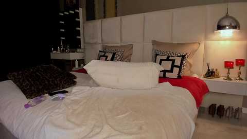 Video of a modern bedroom with a white headboard, white bedding, and leopard-print pillows. A white nightstand holds a lamp, red candles, and shoes. The room is warmly lit with a pendant light.