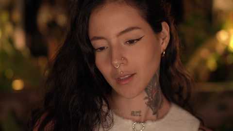 Video of a young woman with long, wavy black hair, wearing a nose ring and tattoos on her neck, smiling softly, set against a blurred, warm-toned background.