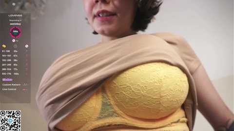 Video of a woman with short, dark hair, wearing a beige top, exposing a yellow lace bra, with a QR code and chat bubbles in the bottom left corner.