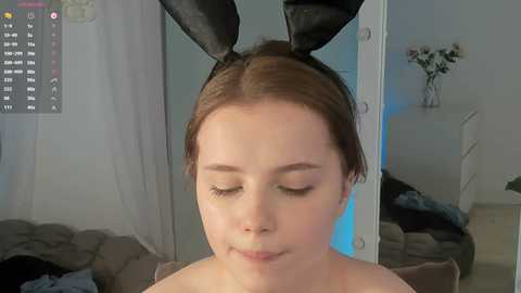 Video of a young Caucasian woman with fair skin, brown hair, and black bunny ears, smiling, in a bedroom with a bed, white walls, and a vase of flowers.