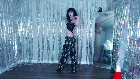 Video of a woman with dark hair and heavy makeup, wearing a black crop top and patterned pants, standing in a room with silver tinsel and a mirror, reflecting a person in the background.