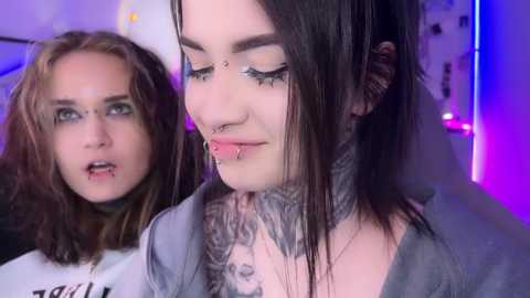 Media: Video of two women in a dimly lit room; one with dark hair, multiple piercings, and tattoos, the other with light brown hair, wearing a white t-shirt.