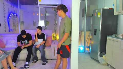 Media: Video of a modern kitchen with a white sofa and four young men, one with curly hair, wearing casual clothes, playing a video game on a phone.
