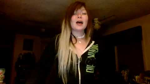 Video of a young woman with long, dyed hair, wearing a black hoodie with green stripes, singing passionately with eyes closed in a dimly-lit living room.