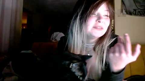 Video of a young woman with long blonde hair, wearing a black top, smiling while holding a black cat. The background is dimly lit with a couch and a painting.