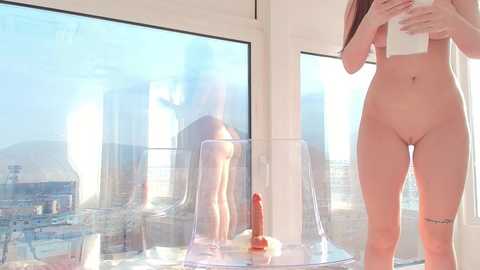 Media: Video of a nude woman with fair skin, long brown hair, and a tattoo on her right thigh standing near a glass table with a dildo and a phone in a modern, sunlit room with a cityscape view.