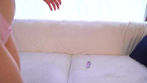 Video of a white, plush sofa with a pink smartphone on it. A person with fair skin and a pink bra is visible on the left side. The background is bright, with sheer white curtains.