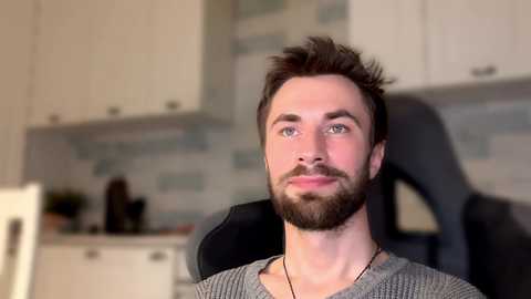 Media: Video of a bearded Caucasian man with short brown hair, wearing a grey sweater, seated in a modern kitchen with white cabinets and blue-tiled backsplash.